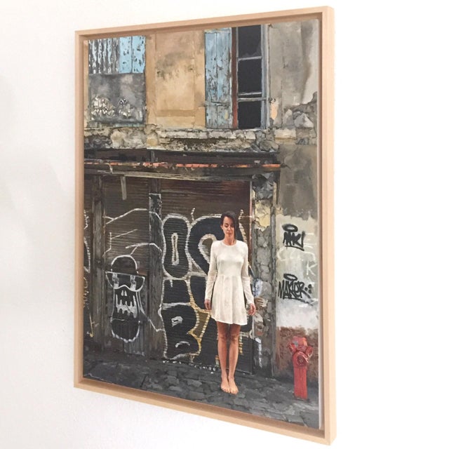 In "Can I Borrow Your Wings," a figure stands barefoot in front of a weathered urban backdrop, characterized by peeling...