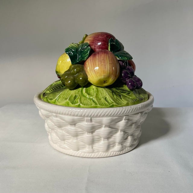 A richly colored tower of faux fruit atop a white basketweave ceramic dish. The bowl features beautifully shaded apples,...