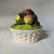 A richly colored tower of faux fruit atop a white basketweave ceramic dish. The bowl features beautifully shaded apples,...