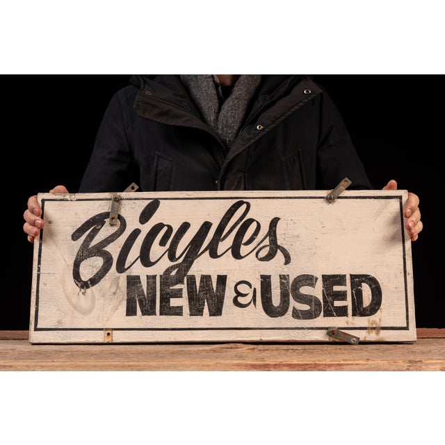 Double-sided painted wood advertising sign reading "Bicycles New & Used" in bold black lettering against a cream ground....