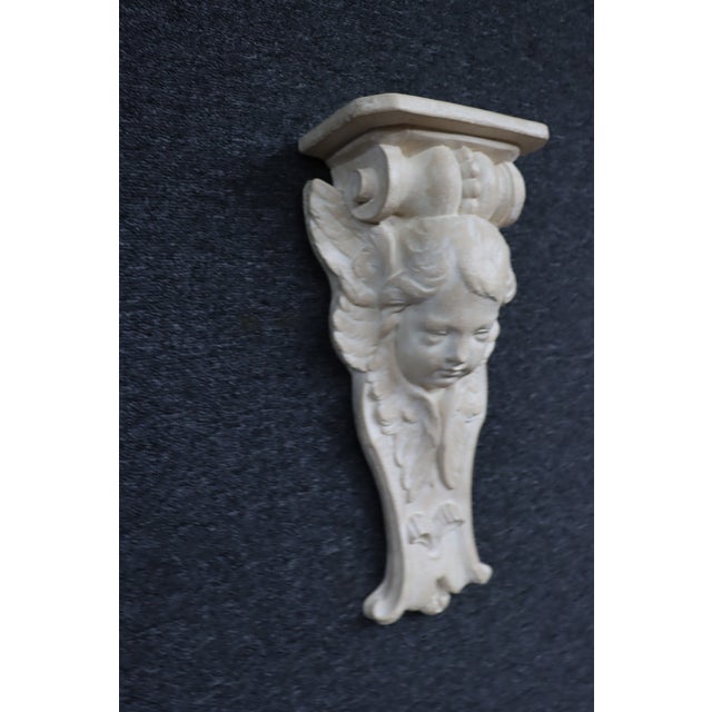 Carved wall bracket featuring a winged cherub supporting a rectangular display shelf. Finished in a faux stone finish with...