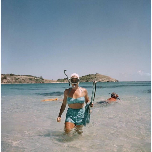 Snorkelling In Antigua by Slim Aarons A woman wearing a scuba diving mask carries a pair of flippers and a harpoon gun,...