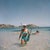 Snorkelling In Antigua by Slim Aarons A woman wearing a scuba diving mask carries a pair of flippers and a harpoon gun,...