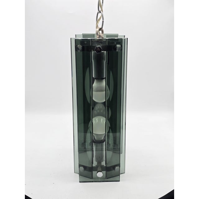 A geometric pendant light composed of thick, green-gray tinted glass panels arranged vertically around a chromed metal...