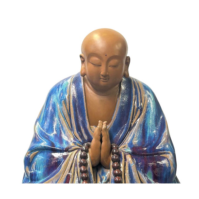 This ceramic sculpture depicts a seated monk or Buddhist figure in a state of deep meditation. The figure is draped in a...