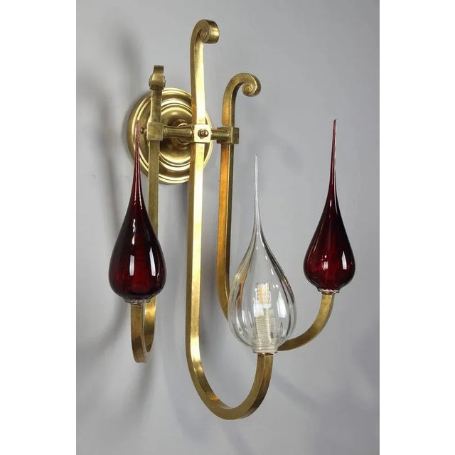 Gilded brass sconce with three scrolled arms of light fixed on the wall with a fluted rosette. Glass and red opaline drop-...