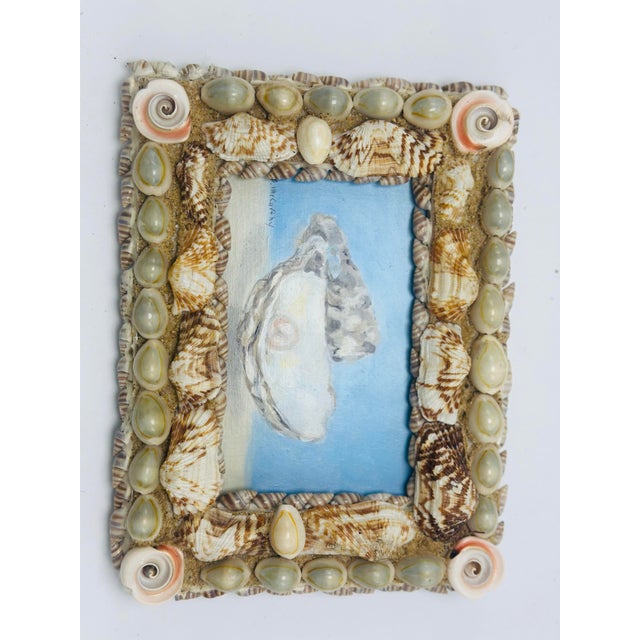 This charming coastal artwork features a small painting of an open oyster shell resting on a sandy shoreline against a...