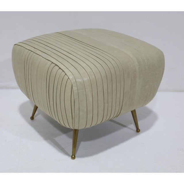 The iconic Kelly Wearstler Souffle Ottoman upholstered in a ivory leather. Brass legs, label underneath. Rushed leather in...