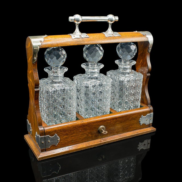 This is a distinguished antique three bottle Tantalus. An English, oak and silver plated drinks caddy with a trio of cut...