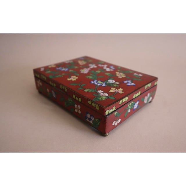 Early 20th century lidded cloisonné rectangular box. Brass construction with hinged lid. Ruby red decorated with colorful...