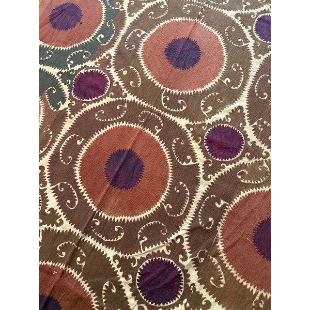 Hand-embroidered Suzani panel from Uzbekistan — rows of terracotta and plum round medallions on a deep brown ground with a...