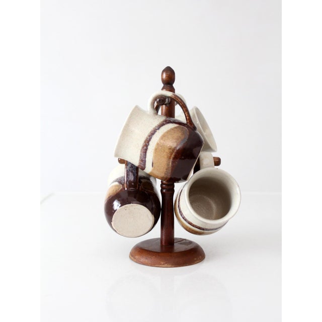 Set of four 1970s handmade studio pottery mugs with a turned wooden stand. Finished in warm cream, brown, and ochre...