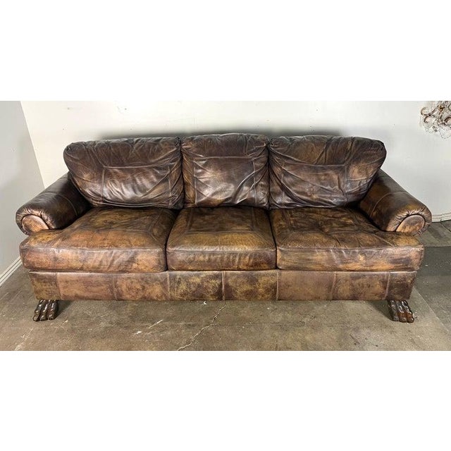 This sofa is a large, three-seat piece with a rustic, vintage style. It is upholstered in a distressed brown leather,...