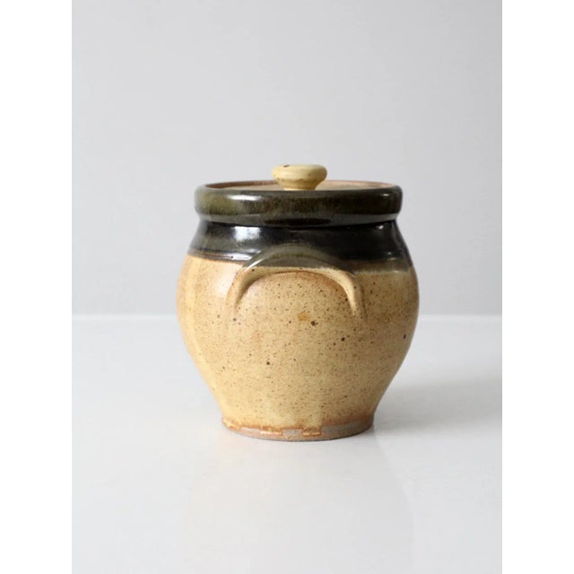 This vintage studio pottery jar brings a warm, organic presence with its speckled tan body and deep olive-brown glazed...