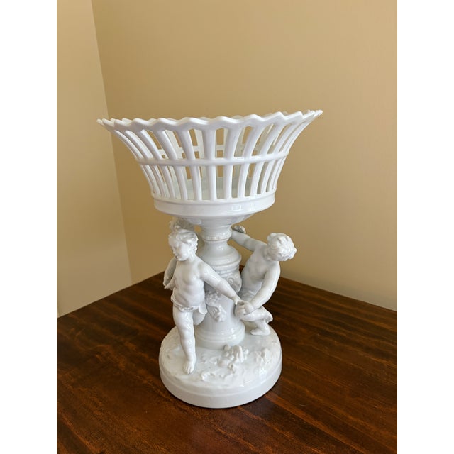 A stunning vintage Neoclassical style porcelain reticulated basket on a pedestal featuring three putti dancing while...