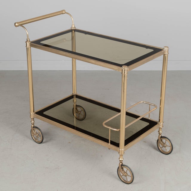 An Italian Hollywood Regency style brass plated bar cart. Smoked glass shelves with dark paned border. Simple bar handle....