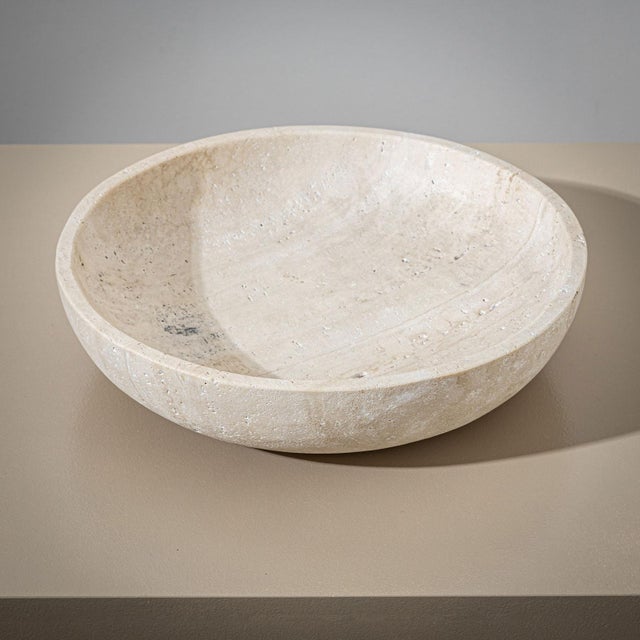 A large, heavy stone bowl crafted from beige travertine, featuring a minimalist design. The bowl is distinguished by its...