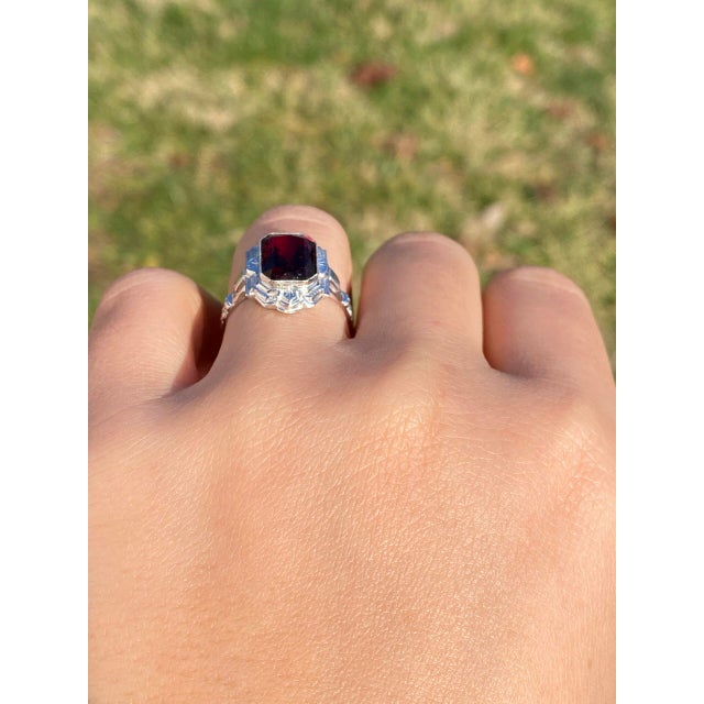 The Trinity River: Antique Art Deco Garnet Ring in White Gold. This exquisite ring showcases a central elongated modified...