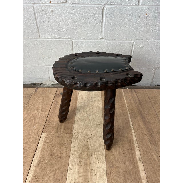 A striking hand-carved rustic tripod stool featuring deeply sculpted organic detailing and a dark leather upholstered seat...
