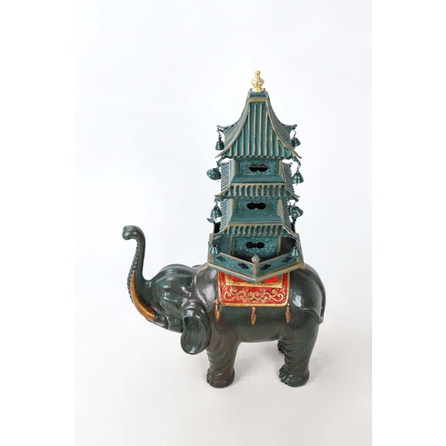 Sculpture of a dark green elephant carrying a pagoda with a lighter shade of green on its back. The addition of a rug...