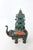 Sculpture of a dark green elephant carrying a pagoda with a lighter shade of green on its back. The addition of a rug...