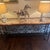 A decorative console table with a hand-forged wrought iron base featuring scrollwork and fleur-de-lis motifs, topped with...