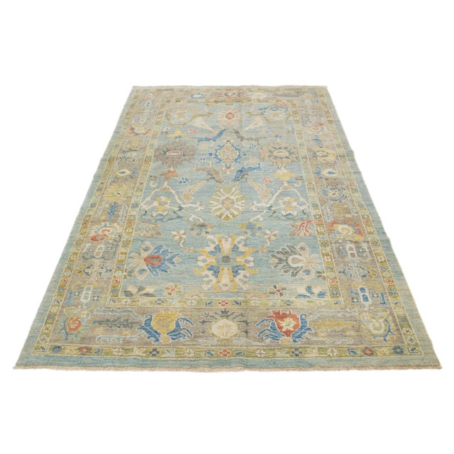 This hand-knotted wool rug features a light blue background with a light brown border and multicolored all-over designs....