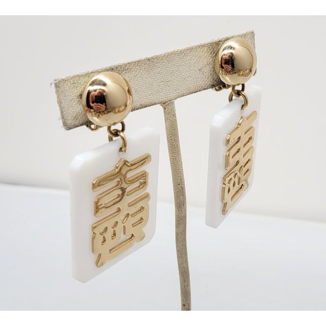 1970s goldtone round domed top with rectangular white resin drop with goldtone asian character overlay clip earrings....