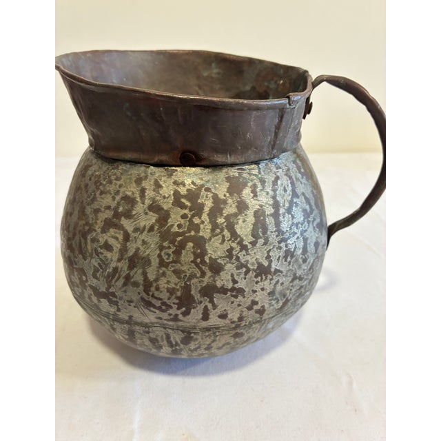 An antique hand hammered copper pitcher with a bulbous riveted body, wide pouring spout, and substantial curved handle....