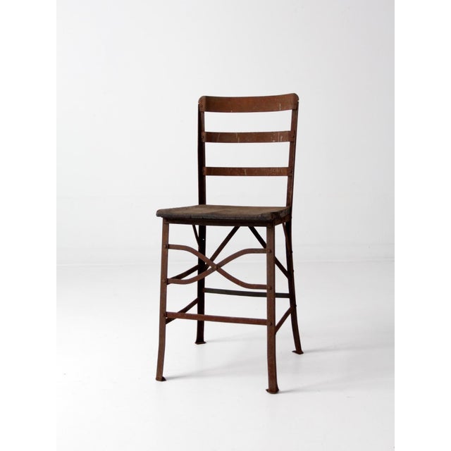 An antique early 20th century chair with a riveted metal frame and wood plank seat. The tall ladder back and gracefully...