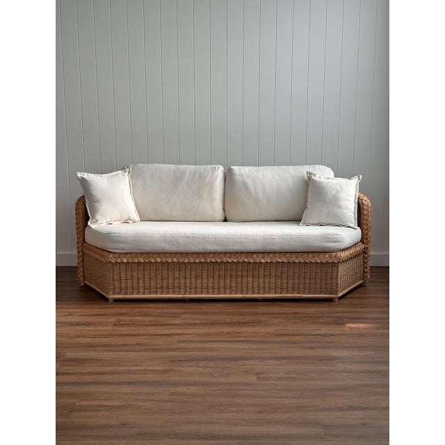 A vintage sofa constructed from braided wicker with a hexagonal frame and low profile, upholstered in white textured...