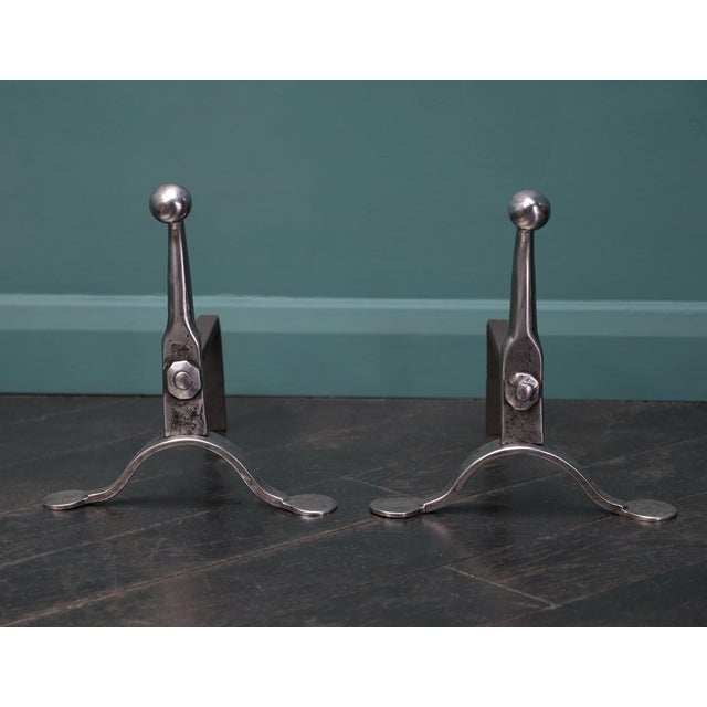 A pair of English mid-19th century polished wrought fire dogs. Wrought uprights with rivets on arched supports with penny...