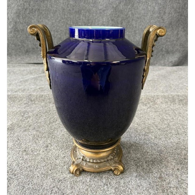 This striking cobalt blue ceramic vase dates to the early 1900s and features ornate brass handles with acanthus leaf...