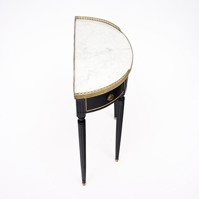 An elegant French Louis XVI style demi-lune console featuring an ebonized wood case, white marble top and refined gilt-...