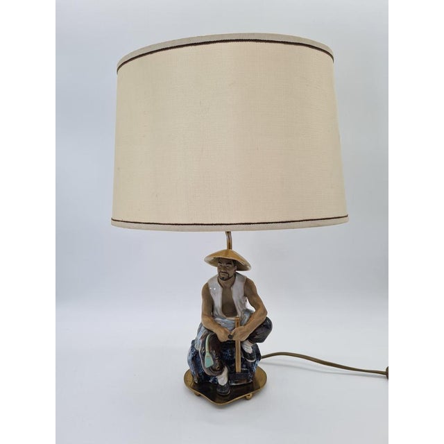 A decorative table lamp from the 1960s–70s featuring a ceramic figurine of a seated fisherman wearing a traditional Asian...