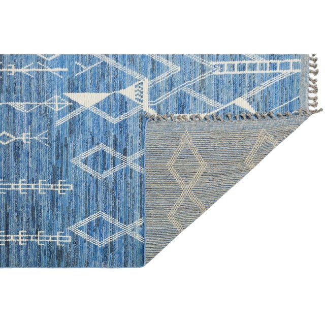 This modern Moroccan-style wool rug features intricate white geometric and symbolic motifs over a rich blue field,...
