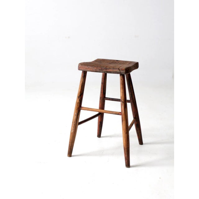 Here is an antique wooden stool. The rustic primitive stool features a saddle seat with splay legs.