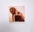 This artwork depicts a figure with long hair, their back turned and head tilted, delicately holding a red apple against...
