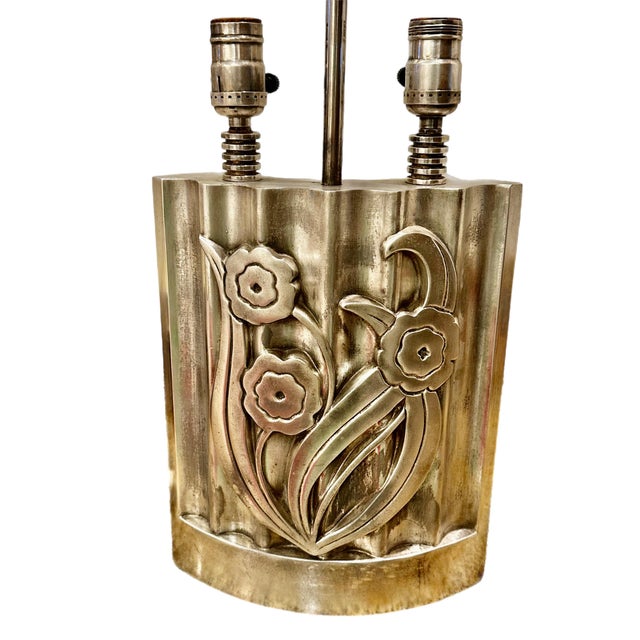 An Art Deco style silver-plated metal table lamp featuring a curved body with a stylized floral relief design on the front...