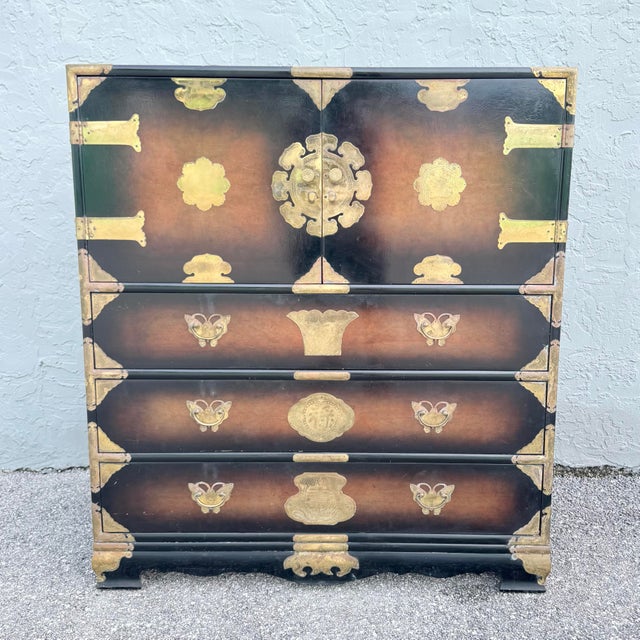 A striking vintage Chinese highboy dresser, richly finished in warm burlwood tones and adorned with elaborate brass...