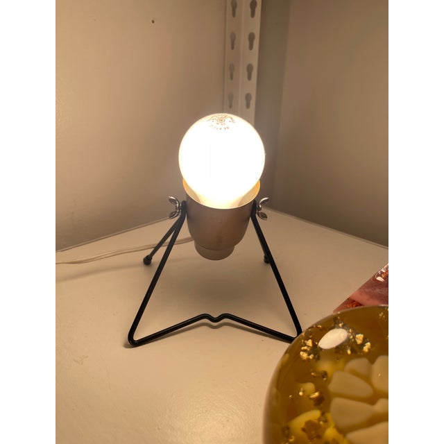 This vintage mid-century modern table lamp features a compact tripod form with an adjustable metal body mounted on a black...