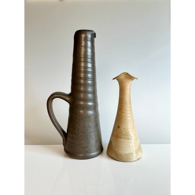 Two slender, tall studio-style ceramic vases made in Germany in the 1980s. The handled vase, in a dark brown with olive...