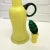 Canary Cased Glass Decanter has a clear base while the decanter has a canary yellow color. It has a green handle attached...
