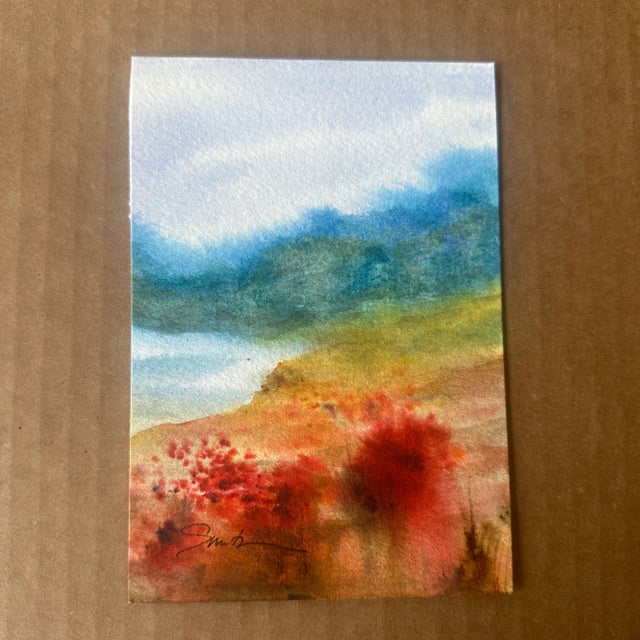 This watercolor painting features a serene landscape with misty blue mountains, a soft sky, and a warm foreground with...