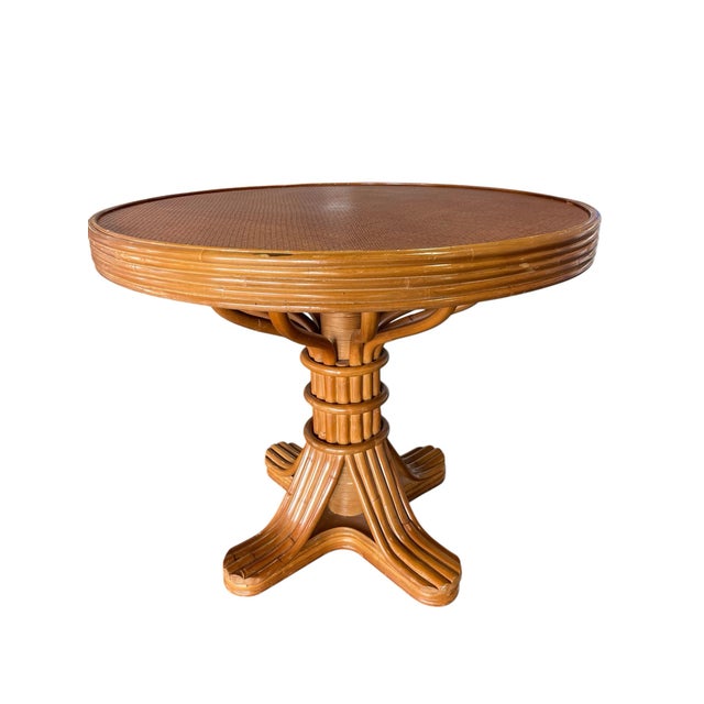 A vintage faux bamboo rattan pedestal dining table featuring a woven inset top and round glass surface. The sculptural...