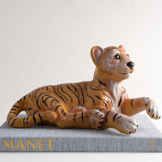Charming ceramic tiger cub sculpture nestled in a laying pose, meticulously hand-painted with rich, lifelike details....