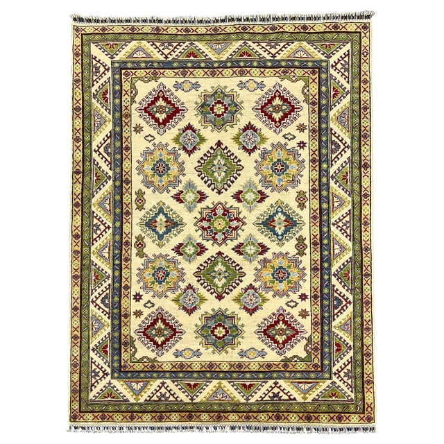 An ivory field displays repeating Kazak-style medallions in burgundy, soft blue, sage green, and gold. Layered patterned...