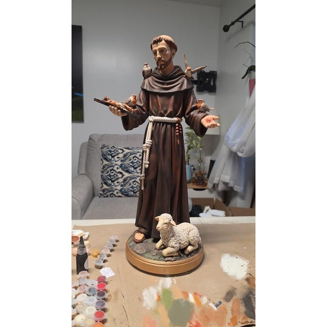 Saint Francis of Assisi is depicted holding a crucifix, accompanied by four birds and a lamb on a circular base. His brown...