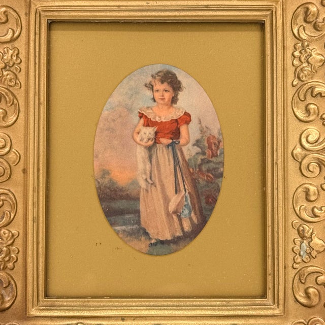 An ornate gold-tone frame surrounds a charming vintage illustration of a young girl holding a white cat. Dressed in a red...