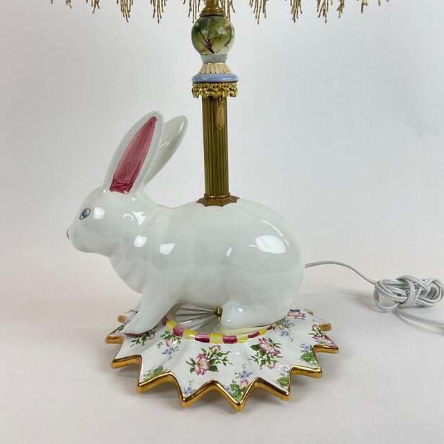 A whimsical and highly collectible MacKenzie-Childs table lamp featuring a sculptural porcelain rabbit base adorned with...